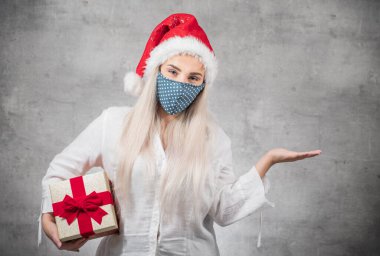 Güzel sarışın kadın Noel Baba şapkası takıyor. Şapka takıyor ve elinde kırmızı hediye kutusu var. Üzerinde kurdele var. Bir Corona - Covid 19 Noel konsepti. Stüdyo çekimi