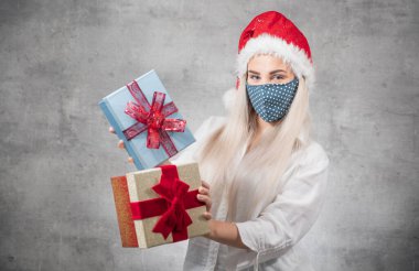 Bir Corona - Covid 19 Noel konsepti. Noel Baba kadın gri arka planda izole edilmiş Noel hediyeleri tutuyor. Kırmızı Noel Baba şapkası takan ve hediye kutusunu tutan mutlu genç kız. Stüdyo çekimi