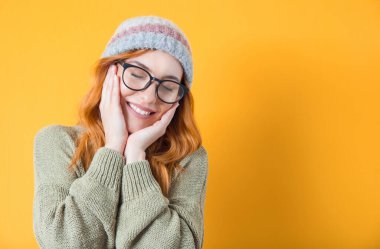 Güzel bir konseptte poz veren, sarı arka planda izole edilmiş, kapalı gözlü tatlı bir kız. Güzel genç bir kadın ellerini yanaklarından ayırmıyor. Stüdyo çekimi