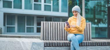 Güzel bir kadın bankta oturuyor ve bir mesaj yolluyor. Açık havada gün ışığı. Moda kızı akıllı telefonuyla göz gezdiriyor. Dışarıda.