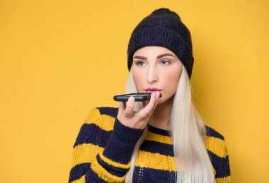Telefondaki genç kadını kapat. Yünlü şapka ve kazak giyen bir manken. Sarı arka planda izole edilmiş. Moda kızı cep telefonunu kullanıyor. Stüdyo çekimi