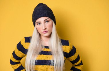 Tarafsız bir yüz ifadesi olan, yün şapka ve kazak giyen, sarı arka planda izole edilmiş, ciddi bir genç kadın. Şirin kız. Stüdyo çekimi