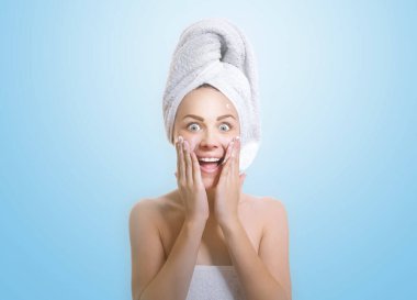 Mutlu kadın yüz kremi sürüyor ya da yanağına nemlendirici sürüyor. Genç kız banyodan sonra, mavi arka planda izole edilmiş.