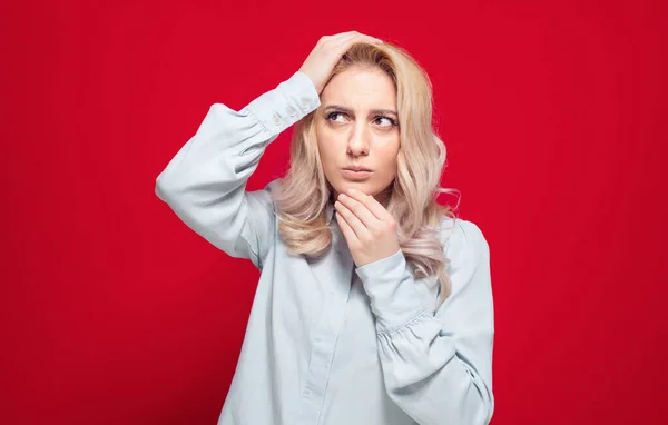 Pensive young woman against white background being in confusion with arms on head, isolated on red background. Studio shot