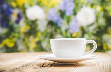 Çay fincanı, yaprakların ve çiçeklerin bulanık arka planının önündeki ahşap masada. Bokeh konsepti - seçici odaklanma. Beyaz çay fincanı. Reklamınız ya da mesajınız için boş bir alan. Kopyalama alanı