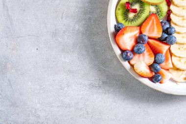 Bir dilim kivi, muz, çilek ve yabanmersini, tabakta meyve salatası. Gri masada sağlıklı bir yemek.