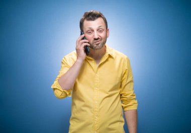 Huysuz adam telefonda mavi arka planda konuşuyor, sarı gömlekli. Baş belası ve akıllı telefon. Stüdyo çekimi