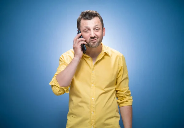 脾气暴躁的男人在电话里用蓝色背景说话 穿着黄色衬衫 疯狂和智能手机 工作室拍摄 — 图库照片
