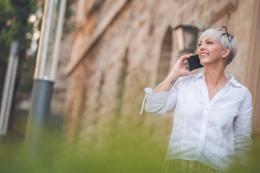 Cep telefonuyla konuşan, kentteki bankta dinlenen orta yaşlı güzel bir kadın. Yetişkin kadın akıllı telefondan konuşuyor. Dışarıda, dışarıda.