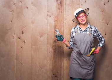 Ahşap arka planda dururken elinde budama makası tutan çekici genç bahçıvan kadın gülümsüyor. Uzayı kopyala Mutlu gülüşlü kadın işçinin yarısı kadar. Stüdyo çekimi.
