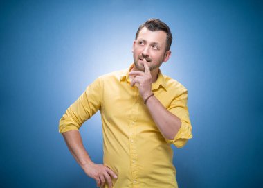 Mavi arka plan üzerinde düşünen, sarı gömlekli genç bir adam. Dalgın adam yukarı bakıyor. Stüdyo çekimi