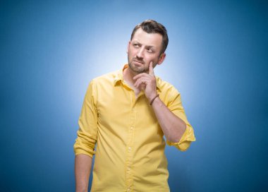 Kurnaz genç adam düşünüyor. İlham arayan zeki insanın belden yukarı portresi, mavi arkaplanda metin için kopya alanı, sarı gömlekli. Düşünceli adam yukarı bakıyor. Stüdyo çekimi