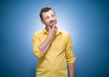 Gülümseyen düşünceler genç adamın düşünceleri. Gülümseyen yüz ifadesi olan esinlenilmiş adamın yarım boy portresi mavi arkaplan üzerindeki metin için kopya alanı, sarı gömlekli. Stüdyo çekimi