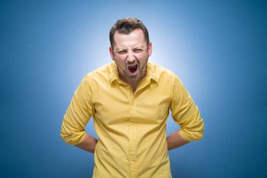 Öfkeli genç adam mavi arka planda çığlık atıyor, sarı gömlekli. Hüsrana uğramış adam. Stüdyo çekimi