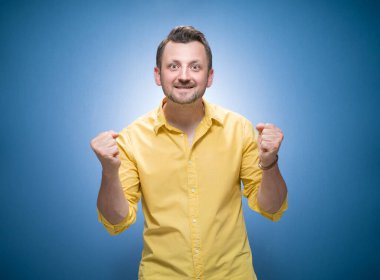 Heyecanlı bir adam ağzı açık bağırıyor ve mavi arka planda yumruklarını sıkmış, sarı gömlekli. Şaşırmış adam başarıyı kutluyor. Stüdyo çekimi