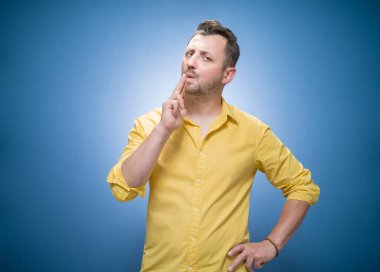 Genç adam ateş etmeye hazırlanıyor, sakallı adam silahını mavi arka planda gösteriyor, sarı gömlek giyiyor. Stüdyo çekimi