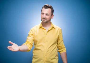 Dost canlısı genç adam mavi arka planda el sıkışmak için elini uzatıyor, sarı gömlekli. Selamlama hareketi konsepti. Stüdyo çekimi