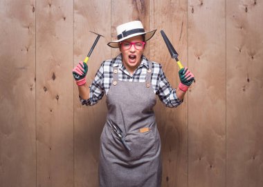 Stüdyoda ahşap arka planda çığlık atarken bahçe aletlerini gösteren öfkeli genç bahçıvan kadın. Hüsrana uğramış kadın çiftçi. Stüdyo çekimi