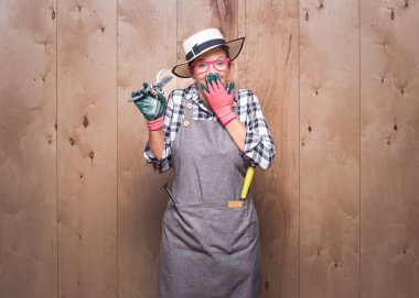 Şaşırmış kadın çiftçi stüdyoda ahşap arka planda kameraya bakıyor. Neşeli genç bahçıvan kadın elinde budama makası ile ağzını eliyle kapatıyor. Stüdyo çekimi