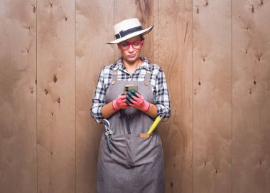 Stüdyoda ahşap arka planda cep telefonunu tutan kafası karışmış genç bahçıvan kadın. Sinir bozucu suratlı, akıllı telefon tutan bir kadın. Baş belası konsept. Stüdyo çekimi