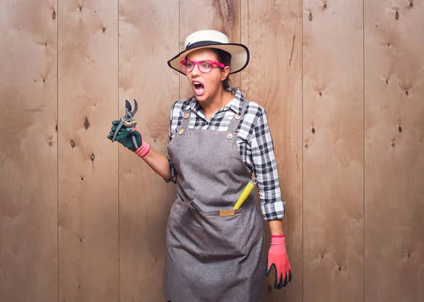 Stüdyoda ahşap arka plana bakarken elinde budama makası tutan, bağıran ve bağıran agresif genç bahçıvan kadın. Stüdyo çekimi