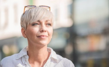 Sarışın, kısa saçlı, güneş gözlüğü takan orta yaşlı güzel bir kadın. Beyaz yaz gömleği giymiş kadın profili. Açık havada, şehrin bulanık sokaklarında..