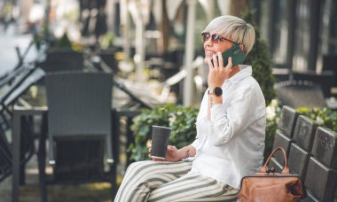 Cep telefonundaki kadın dışarıda, kentteki bankta konuşuyor ve dinleniyor. Akıllı telefondan konuşan yetişkin bir kadının profili ya da yan görüntüsü.