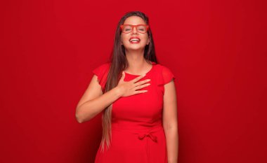 Kendinden geçmiş genç bir kadın elini göğsüne koyar, kırmızı arka planda izole olur. Memnun güzel kız. Stüdyo çekimi