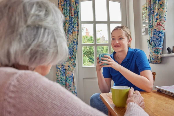 Female Home Help Having Cup Of Tea With Lonely Senior Woman In Kitchen