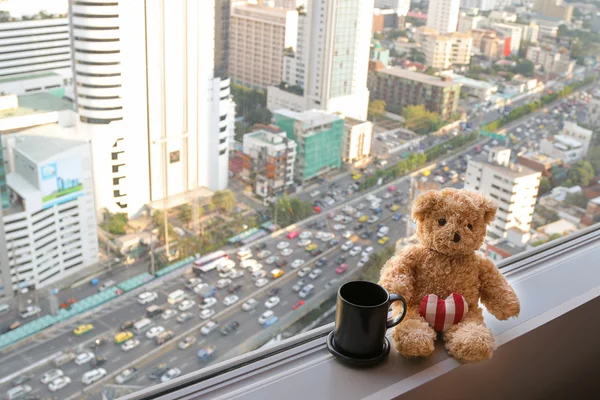 A bear sitting next to the window with a coffee mug and blurred traffic congestion — Stock Photo, Image