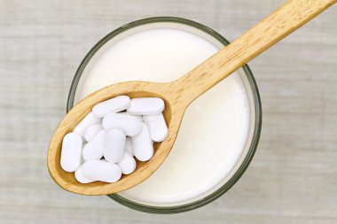 Wooden spoon of white Calcium carbonate tablets above a glass of fresh milk clipart