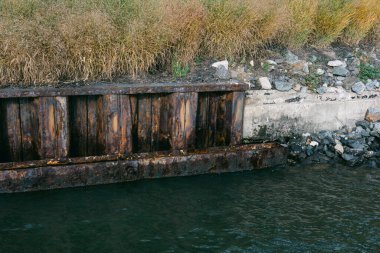 Köy ahşap rıhtımı, çimenler ve kayalık kıyı şeritleriyle çevrili, kısmen sakin sulara gömülü..