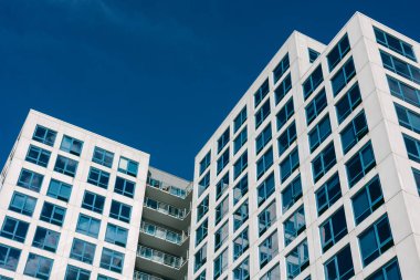 Büyük pencereli iki modern apartman, parlak mavi bir gökyüzüne karşı duruyor, şehri simgeliyor..