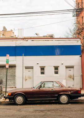 Şehrin bulutlu bir gününde, eski bir bordo araba kaldırım kenarında mavi ve beyaz bir binanın yanına park edilmiştir..