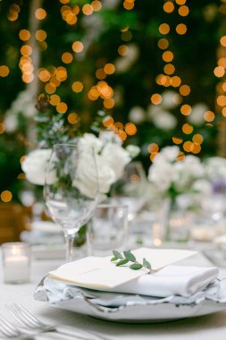 Güzel bir açık hava masasında beyaz çiçekler, parıldayan ışıklar ve zarif bir menü kartı sergilenir..