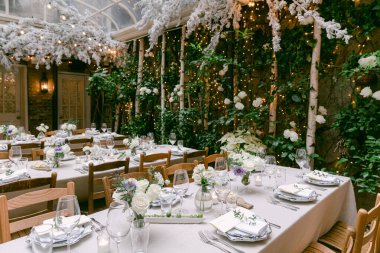 Güzel dekore edilmiş kapalı bir bahçede, çiçekli ve sıcak ışıklandırmalı resmi bir yemek için hazırlanmış şık masalar..