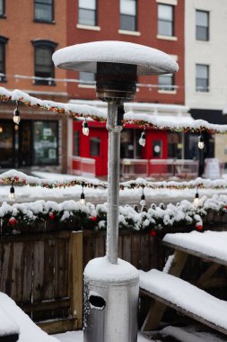 Bir açık hava ısıtıcısı, kışın etrafı tatil süslemeleri ve ışıklarla çevrili bir şehir restoranında karın içinde duruyor..