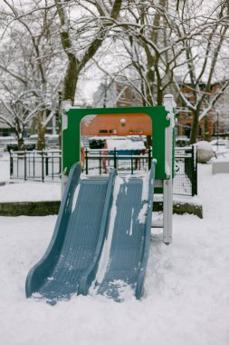 Oyun parkında bir kış günü ağaçlarla çevrili iki mavi slayt yer alır..