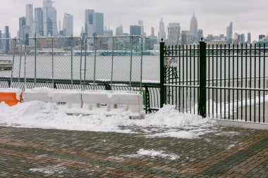 Önünde bir çitle yerde kar var ve bulutlu bir günde arka planda bir şehir silueti..