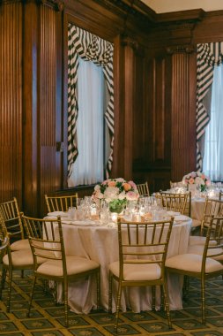 Çiçekler ve mumlarla süslenmiş masalar resmi bir alanı uzun tahta duvarlar ve çizgili perdelerle doldurur..