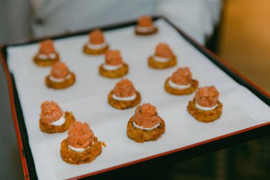 Tepside küçük, yuvarlak aperatifler var. Üstüne pembe karışım ve beyaz serpiştirilmiş. Misafirler sosyal etkinliklerde yemek yerler..