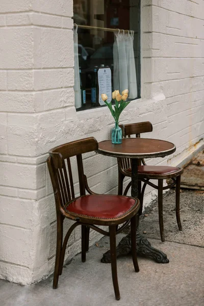 İki ahşap sandalye ve bir vazo çiçekli küçük bir masa bir duvarın yakınındaki kafenin önüne kurulur..