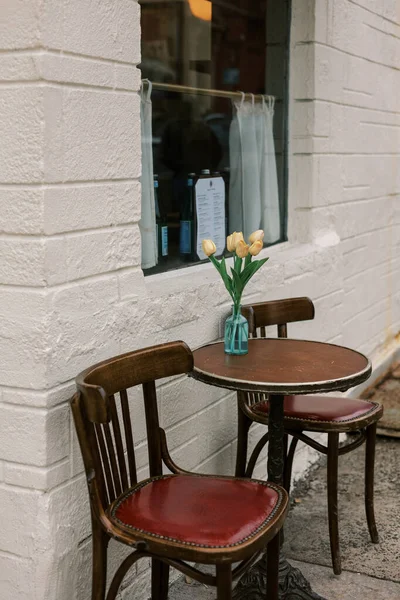 İki ahşap sandalye ve küçük bir masa bir kafenin önünde vazoda çiçeklerle oturup, basit bir sahne yaratıyorlar..