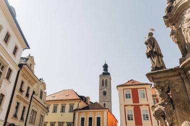 Uzun saat kulesi şehir meydanında duruyor. Binalar ve bir heykel. Gün ortasında canlı bir atmosfer sergiliyor..