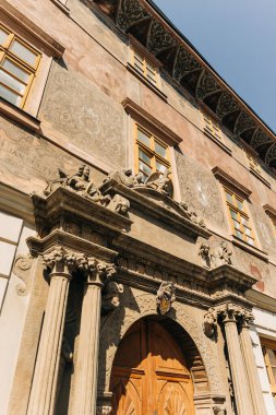 Tarihi bir binanın girişi, parlak gökyüzü ve güneş ışığı altında detaylı taş oymalar ve ahşap kapılar gösteriyor..