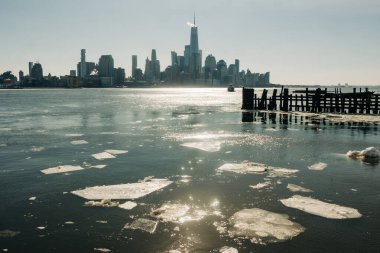 Sahne New York şehrinin ufuk çizgisini gösteriyor. Su buzlu ve güneş ışığı yüzeyden yansıyor..