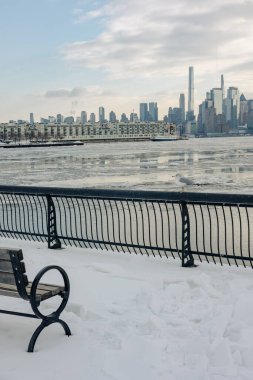 Nehrin yanındaki karların üzerinde bir bank oturuyor. Kış günü ufuk çizgisinde yüksek binalar var..