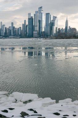 Gökdelenler, kışın nehre bakan kentsel bir arazide buzlu suyun yanında görünür yansımalarla dururlar..