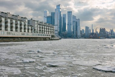 Bulutlu bir gökyüzüne karşı yükselen yüksek binalarla, buzla kaplı bir nehir şehrin içinden akar..