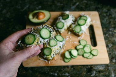 Salatalık dilimleri avokadoyla birlikte bir tabağa konur. Bir el kesme tahtasının üzerinde bir parça tutuyor..